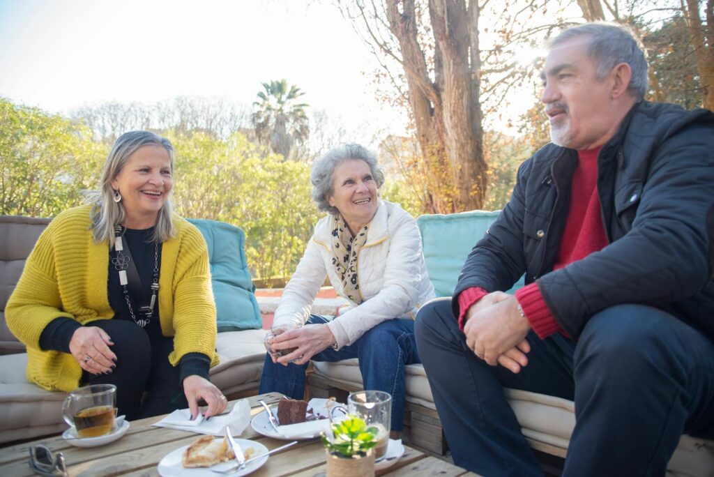 Three senior friends enjoying coffee and cake outdoors in a sunny park setting in Edmonton Retirement Community